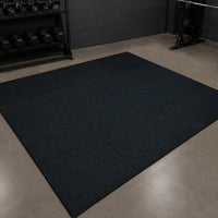 A large black rubber gym floor mat made of interlocking square tiles, shown in a gym setting on a concrete floor with weight racks in the background.